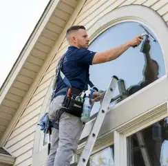 Professional window cleaner on a ladder using a squeegee to clean an exterior arched window, Glendale, AZ.